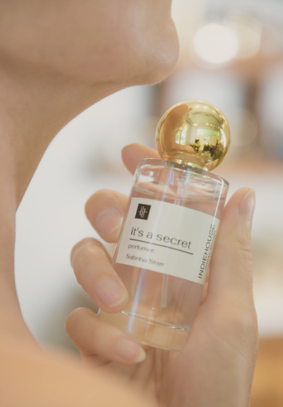 Person holding Indiehouse perfume bottle labeled "It's a Secret" with a gold cap, promoting custom scent creation in Atlanta.